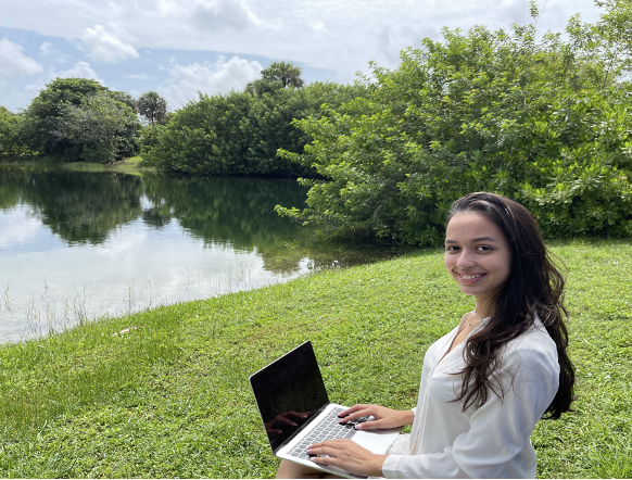Woman working on a laptop as she sits in front of a lake.