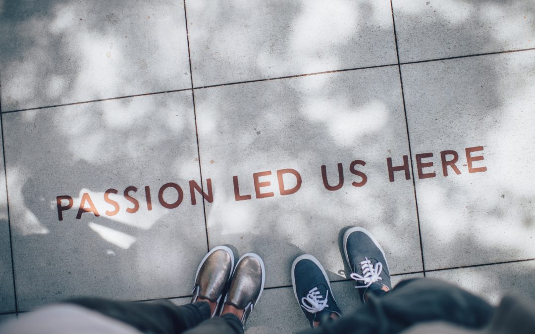 Text "Passion Led Us Here" painted on a sidewalk with two people's feet hovering under it.