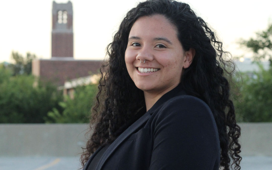 A woman wearing a black blazer with a campus backdrop smiling at the camera.