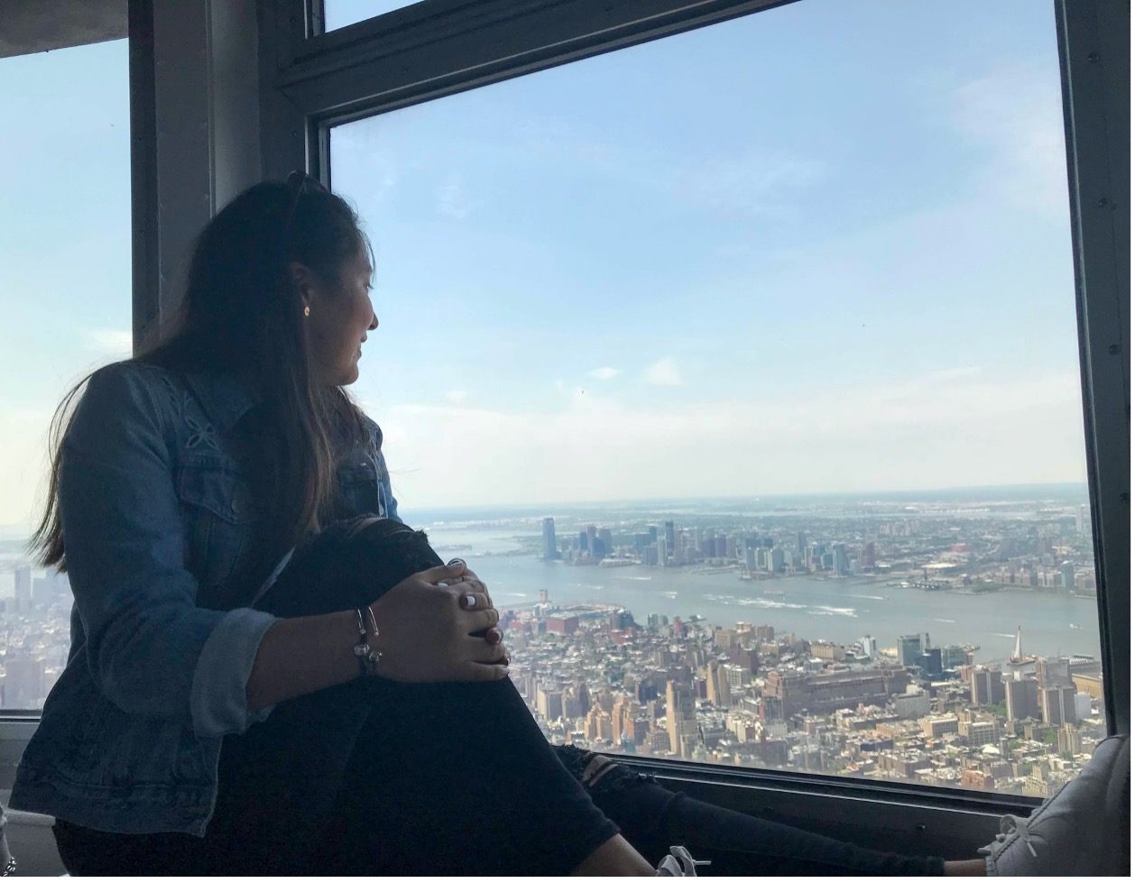 A Latinx woman sitting on a window ledge looking out onto an aerial view of a city.